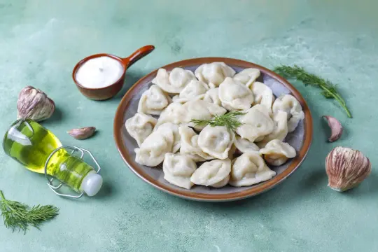Talerz pełen pysznych, domowych pierogów, obok czosnek, koperek i buteleczka oliwy. Różne rodzaje pierogów zachęcają do spróbowania.