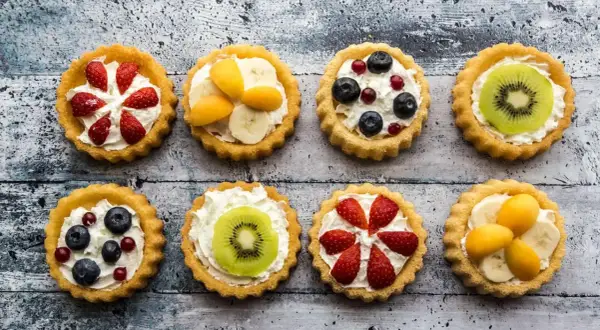 Osiem kolorowych tartaletek z owocami: truskawkami, borówkami, kiwi, bananami i morelami na kremowym nadzieniu.