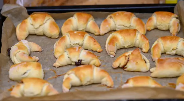 Szybkie rogaliki kruche, świeżo upieczone, leżą na blasze wyłożonej papierem do pieczenia. Niektóre z nich mają nadzienie.