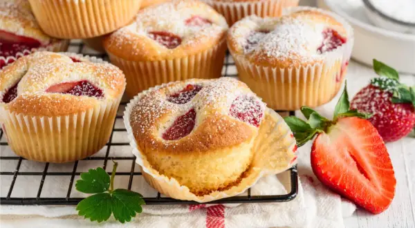 Puszyste muffiny z truskawkami, posypane cukrem pudrem, prezentują się apetycznie na kratce. Obok leżą świeże truskawki.