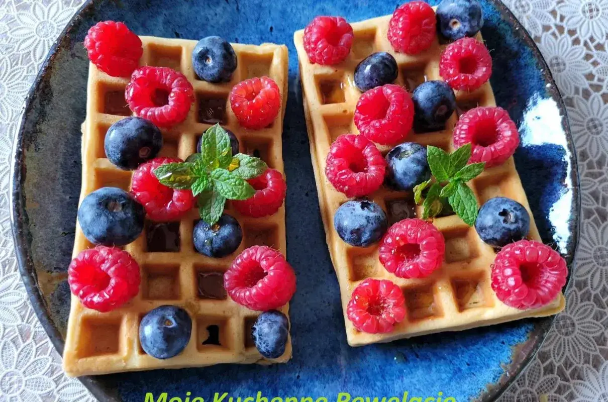 Dwa keto gofry z mascarpone, malinami, borówkami i miętą na niebieskim talerzu.